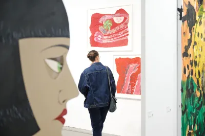 A woman looks at two prints in an art fair booth, with other works seen in the foreground. 