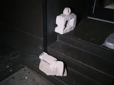 A photo shows two small sculptures in gray stone sitting on black metal steps against a black wall